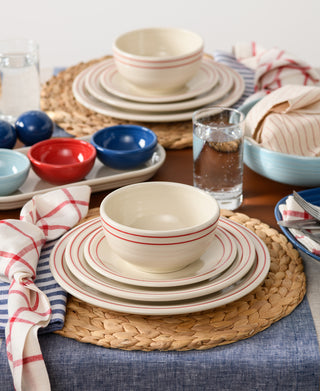 A table set with the Fiesta Americana Scarlet Band 3-Piece Place Setting, woven placemats, blue and red bowls, striped napkins, glasses of water, and a blue cloth underneath creates a casual and colorful dining scene.