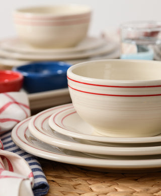 A close-up of the Fiesta Americana Scarlet Band 3-Piece Place Setting shows cream-colored ceramic plates and a bowl with red stripes, set on a woven placemat, with colorful napkins and tableware in the background.