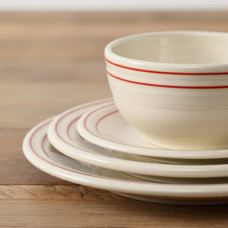 A close-up of the Fiesta Americana Scarlet Band Classic Bistro 3-Piece Place Setting, Service For 1, features stacked cream-colored dishes and bowls accented with red stripes. Surrounded by striped napkins and small colorful bowls on a woven placemat, this proudly made in the USA set brings vibrant charm to any table setting.