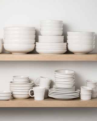 Two wooden shelves display neatly stacked white ceramic dishes, bowls, plates, and mugs against a plain white wall. The dinnerware is arranged in organized, evenly spaced piles.