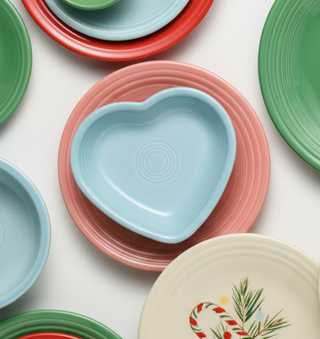 A collection of ceramic plates and bowls in festive holiday colors, including a light blue heart-shaped bowl, a pink plate, green plates, and a cream plate with a candy cane and pine design, arranged on a white background.