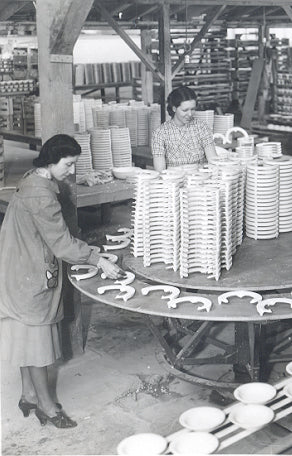 Two women work in a ceramics factory arranging and inspecting stacked plates and dishware on a round table, surrounded by shelves filled with pottery.