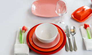 A table setting with red and pink plates, a white bowl, silver cutlery, a wine glass, a red butter dish, and napkins each holding a red tulip on a white tablecloth.