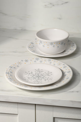A set of white dinnerware with blue and gold snowflake patterns, including two plates and a bowl, arranged on a white marble countertop.