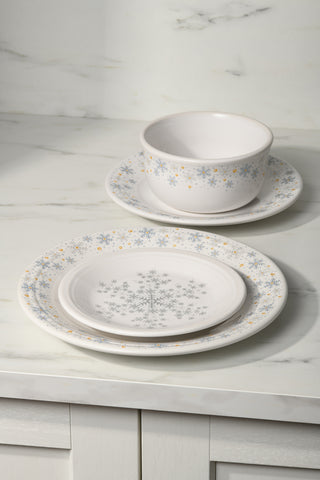 A Fiesta (FTC) White Snowflake Classic Rim 7 1/4 Inch Salad Plate sits on a marble countertop, its white ceramic surface decorated with elegant blue and gold snowflake patterns from the festive collection.