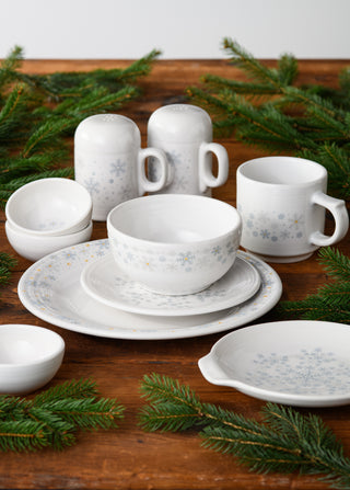 The Fiesta White Snowflakes 16oz Stackable Mug, crafted with lead-safe china, is featured alongside coordinating plates, bowls, and shakers on a wooden table decorated with green pine branches for a festive touch.