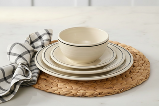 A stack of cream-colored dishes and the Fiesta Black Band Bistro Coupe 5 1/2 Inch Cereal Bowl, featuring sleek black rims, sit on a woven placemat next to a folded black-and-white checkered napkin atop a marble surface.