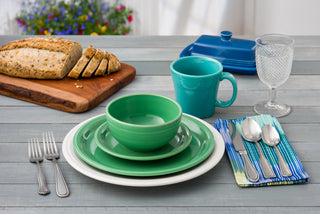 A table setting with green dishes, a Fiesta 15 OZ Tapered Mug by Fiesta made in the USA, silverware on a striped napkin, a glass of water, a blue butter dish, sliced bread on a wooden board, and flowers in the background.