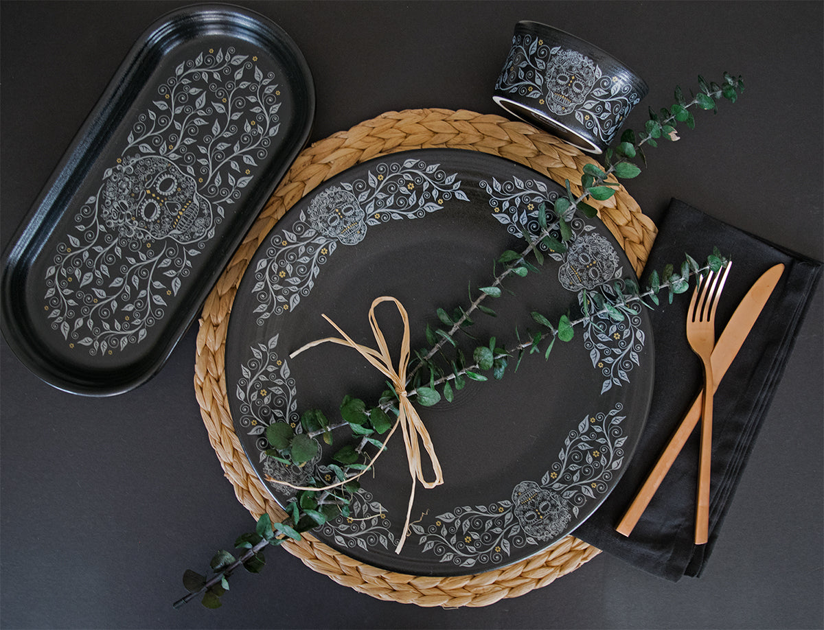 Balck tablesetting with black SKULL AND VINE patterend bread plate, ramekin and dinner lpate. On top of the plate is tied ecalyptus. Also on the table is black napkin and gold flatware