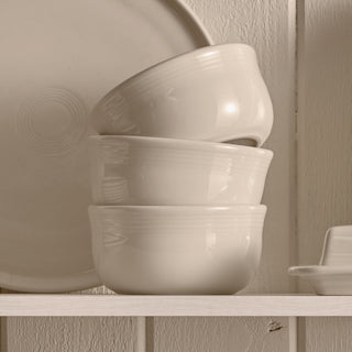 Three Fiesta All-Purpose 6 Inch Gusto Bowls (28 OZ) in white are stacked on a shelf before a large plate and beside a small dish, all made of lead-free ceramic and set against a white wooden wall.