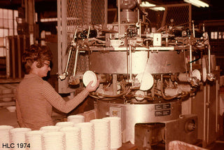 A woman operates a large industrial machine, placing white ceramic plates onto it. Stacks of plates are on a table nearby. The image appears to be from a factory setting in 1974.