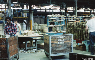 Workers process clay in a large industrial factory. Wooden bins labeled SCRAP CLAY ONLY are in the foreground, and stacks of clay pieces and machinery fill the busy workspace. The image is dated HLC 1986.