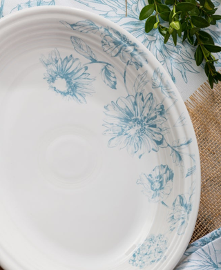 The Fiesta Botanical Floral Sky 11 5/8 Inch Medium Oval Serving Platter, made in the USA, features light blue floral designs on white ceramic and is displayed on a blue and white patterned cloth with burlap and green leafy branches nearby.