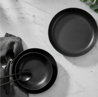 A stylish table showcases the Fiesta Coupe Bowl 3-Piece Place Setting in Foundry, with two black vitrified china plates and a matching coupe bowl on marble. A gray napkin holds black cutlery to the left, while green leaves peek in from the top right.