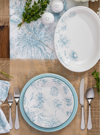 A rustic table is set with a Fiesta Botanical Floral Sky 11 5/8" Medium Oval Serving Platter, silver utensils, a twine-tied napkin, greenery, and white-blue flowers on burlap placemats over wood.