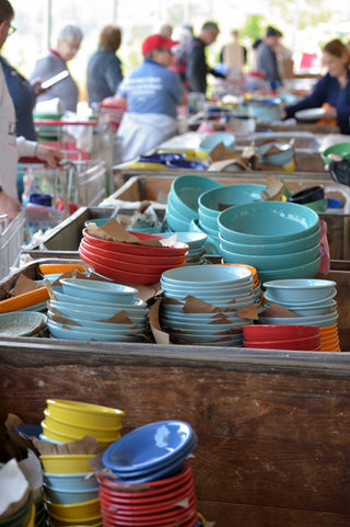 Shoppers browsing bins filled with discounted ceramic dinnerware in various colors, including blue, red, yellow, and turquoise, at an outdoor tent sale.