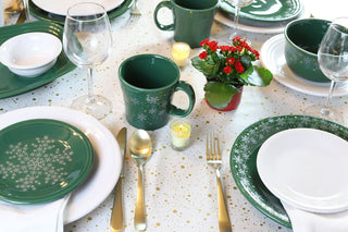 A festive table setting with green plates and mugs featuring white snowflake designs, white plates, gold utensils, clear glasses, small candles, and a red flowering plant centerpiece on a white and gold dotted tablecloth.