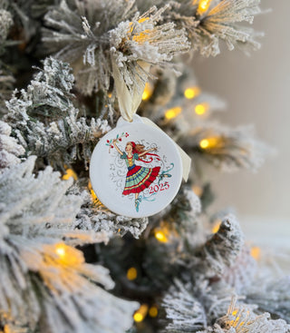 The Fiesta Nutcracker Ballerina 2025 Ornament, made in the USA, displays a vibrant dancing ballerina and the year 2025. It hangs on a snow-dusted tree lit with warm yellow lights and frosted branches.